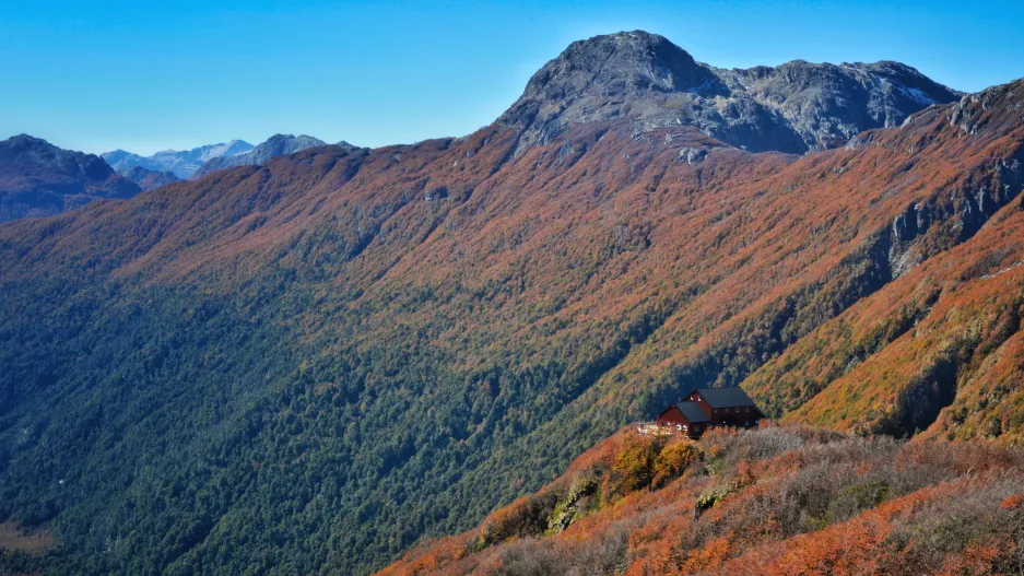 una ladera con tonos verdes abajo y naranjas arriba antes de llegar a la cumbre de piedra. Abajo a la derecha aparece el refugio Rocca con chapas rojas y techo negro