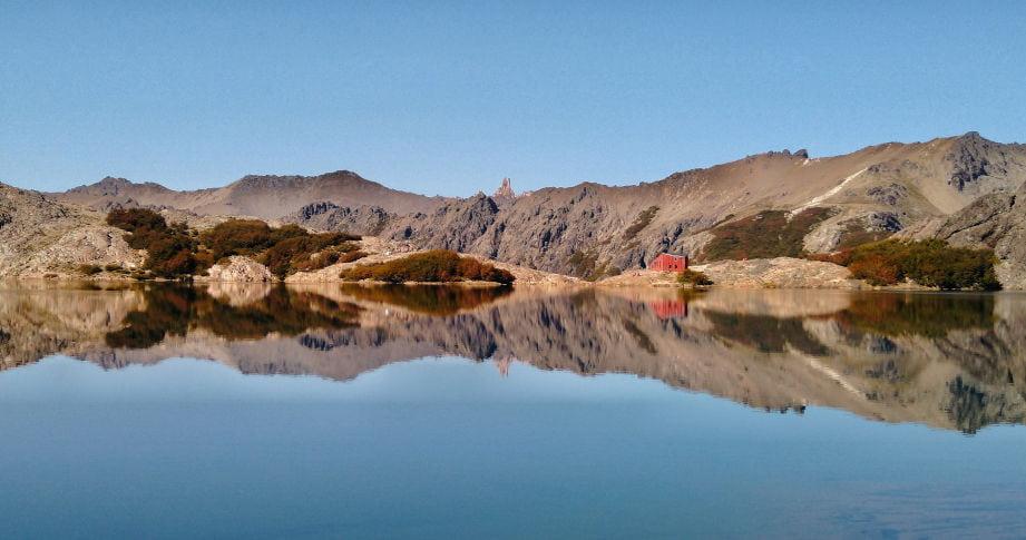 imagen de un espejo de agua en calma con un refugio de montaña al fondo y el cielo celeste
