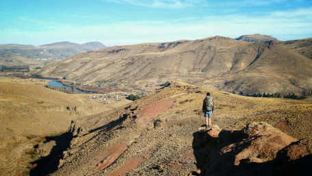De espaldas sobre una roca, una persona sola observando el paisaje de la estepa en Villa Llanquín. El río Limay de celeste fluye entre el suelo desértico.