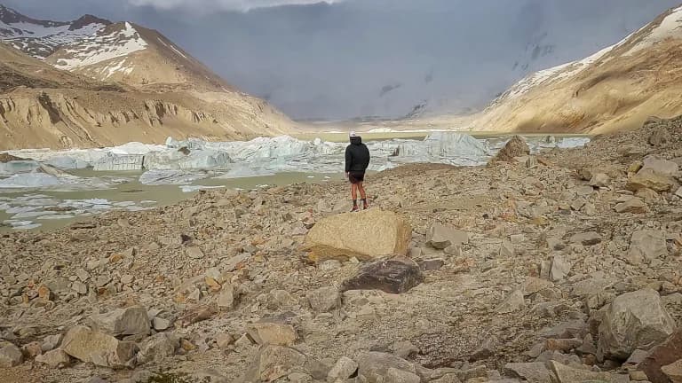 una persona parada sobre una roca con la laguna de los témpanos al fondo. La laguna es de color gris lechoso y hay bloques de témpanos de hielo.